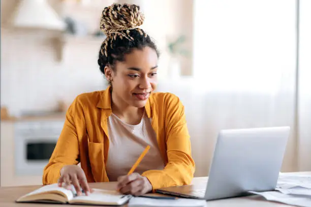 Photo of Focused cute stylish african american female student with afro dreadlocks, studying remotely from home, using a laptop, taking notes on notepad during online lesson, e-learning concept, smiling