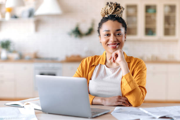 soddisfatta giovane donna afroamericana elegante, freelance, studentessa o agente immobiliare, seduta alla sua scrivania all'ufficio di casa, guardando la telecamera e sorridendo piacevolmente - laptop using laptop studying working at home foto e immagini stock