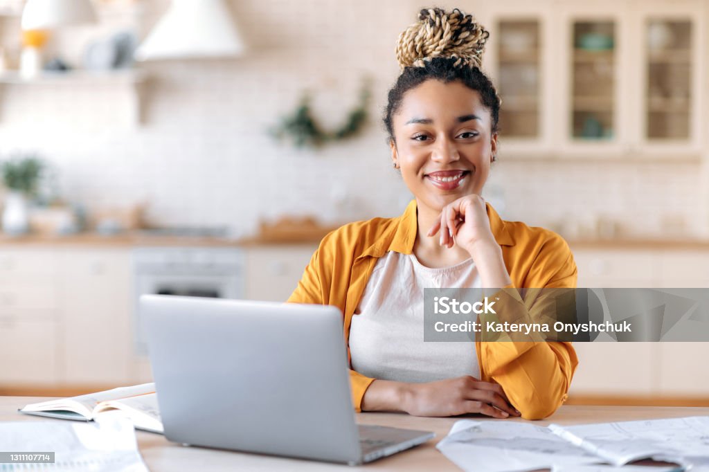 Satisfied good looking young African American stylish woman, freelancer, student or real estate agent, sitting at her desk at home office, looking at the camera and smiling pleasantly Women Stock Photo