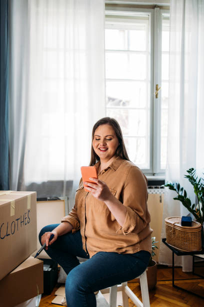 istock Moving Out: Smiling Plus Size Woman Sitting next to a Cardboard Box while Texting on her Smartphone 1311108307