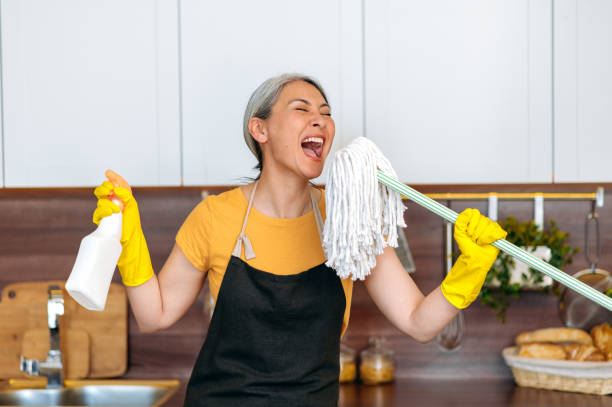 happy funny hardworking mature gray-haired asian female cleaning worker or housewife in gloves and apron, taking a break, dancing in the kitchen with detergent and mop, singing her favorite song - spring cleaning women cleaning dancing imagens e fotografias de stock