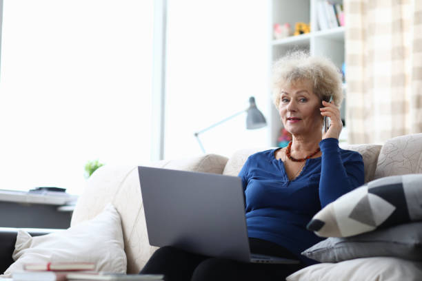 istock Elderly woman talking on smartphone holds laptop while sitting on couch 1311109906