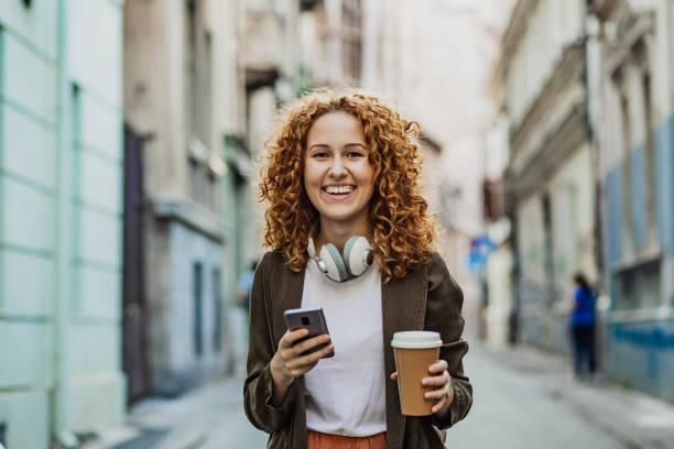 istock Young redhead woman is walking on the street and using mobile phone while holding reusable coffee cup 1311111872