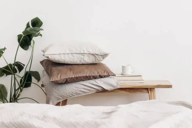 Photo of Trendy Scandinavian interior still life. Velvet and linen cushions on vintage wooden bench, table. Cup of coffee on pile of books and monstera potted plant. White wall background. Blurred bed