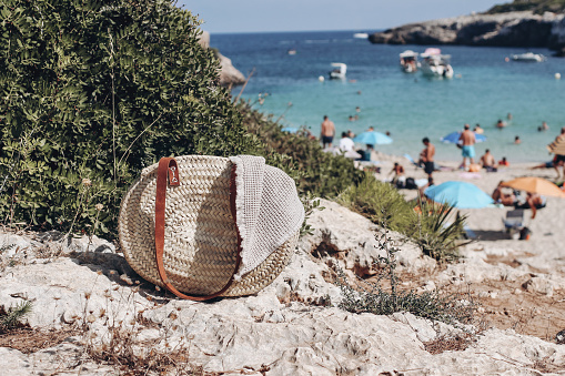 Summer travel, vacation still life. French straw basket with beige towel. Blurred background with sea, boats and swimmers.Sunbathing people, tourists. Selective focus. Mallorca island beach.