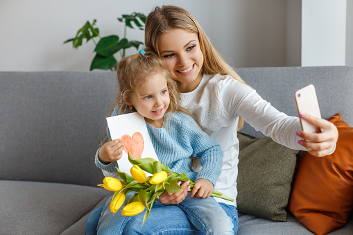 International women's and mother's day. Little cute daughter with bouquet of yellow flowers and postcard congratulates smiling mom. Small girl make selfie with mommy. Happy family holidays at home