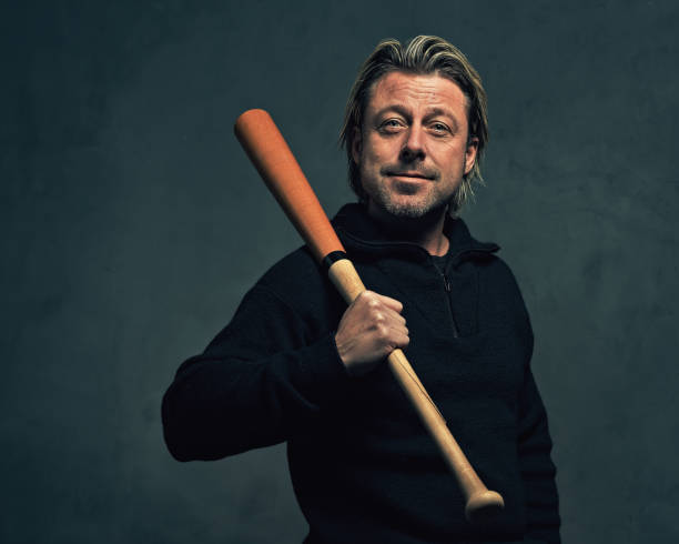istock Smiling blond man with stubble beard in black wool sweater holding a baseball bat in front of a dark grey wall. 1311116997