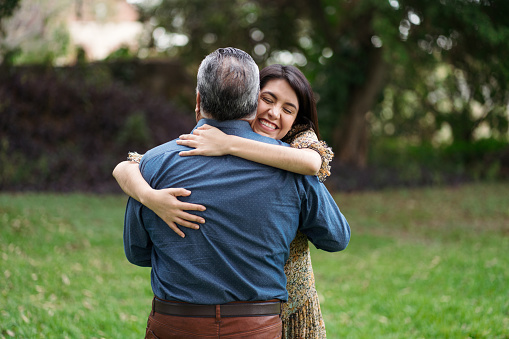 A happy young adult daughter embracing her father outside and smiling with eyes closed.