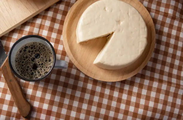 Breakfast table in Brazil with breads, cheese, cup of coffee and accessories on a brown and beige checkered tablecloth, dark background, top view.
