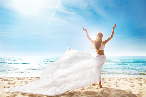 Happy Freedom Woman on Beach Enjoying Sun Open Arms Outstretched. Rear View of Girl Raised Hands fluttering White Dress on Wind. Summer Holiday Tropical Vacations Travel. Carefree Relax Rest