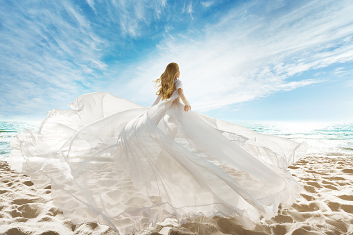 Woman on Beach in White Dress flying on Wind. Summer Holiday Travel. Tropic Vacations. Sunny Beach, Sand Coastline, Sea and Sunshine Sky