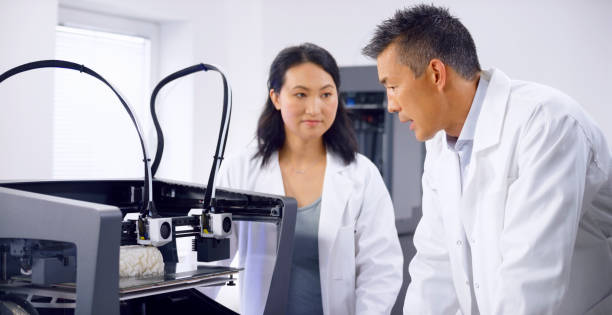 Male and female biomedical engineers talking while looking at 3D printer Male and female biomedical engineers talking about 3D brain model while standing near 3D printer. 3d printing lab stock pictures, royalty-free photos & images