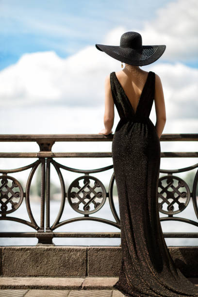 Woman Back Side View in Hat and Evening Dress Outdoor. Fashion Model Rear View looking away. Luxury Lady in long Gown at Promenade looking at Sky Woman Back Side View in Hat and Evening Dress Outdoor. Fashion Model Rear View looking away. Luxury Elegant Lady in long Gown at Promenade Fence looking at Sky evening gown stock pictures, royalty-free photos & images