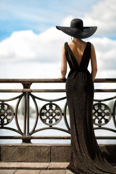 woman back side view in hat and evening dress outdoor. fashion model rear view looking away. luxury lady in long gown at promenade looking at sky - luxury glamour dress caucasian imagens e fotografias de stock