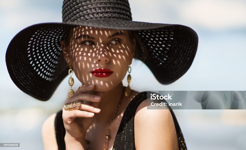 Woman in Hat Portrait. Fashion Luxury Model in Black Summer Hat with Make up and Golden Jewelry. Close up Beauty Face over Sky Background Woman in Hat Portrait Closeup. Fashion Luxury Model in Black Summer Hat with Red Lips Make up and Golden Jewelry. Close up Beauty Face over Sky Background Women Stock Photo