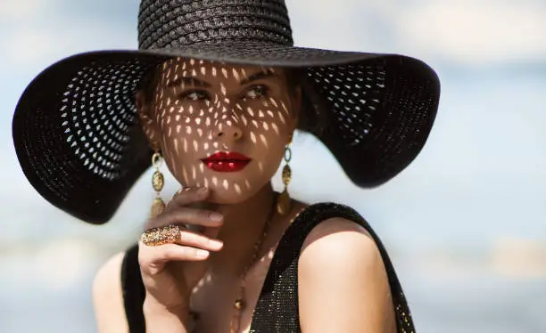 Woman in Hat Portrait Closeup. Fashion Luxury Model in Black Summer Hat with Red Lips Make up and Golden Jewelry. Close up Beauty Face over Sky Background