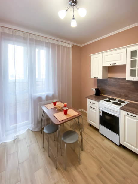 istock Interior of a beige kitchen with a white kitchen set and a stove, a table with utensils and a window with a balcony, covered with white tulle 1311137086