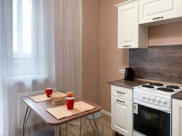 istock Interior of a beige kitchen with a white kitchen set and a stove, a table with utensils and a window with a balcony, covered with white tulle 1311137199