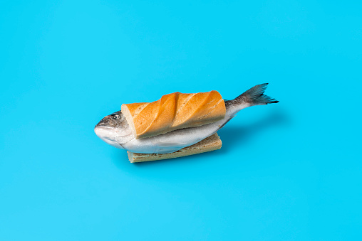 Whole raw fish between slices of bread on a blue colored table. Eating raw fish concept with a frozen dorado fish in a sandwich