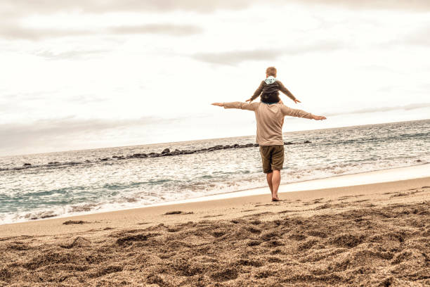 Surfer dad, and his two year old son are holding hands and both carrying their surfboards while walking down the beach
