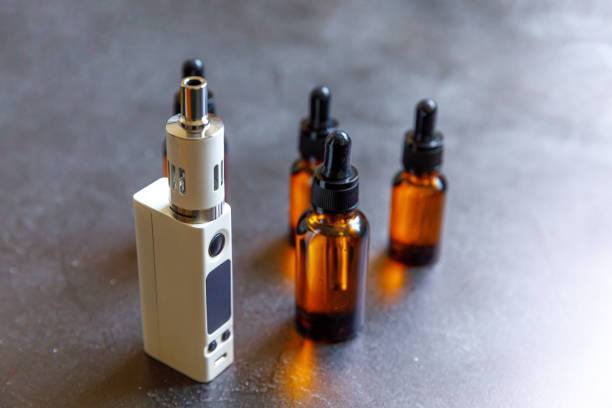 istock Vaping device e-cigarette electronic cigarette and liquid bottles on dark black stone shale background. Vape device for alternative smoking. Vaping shop concept. Gadget for vaper. Vaping accessories 1311142682