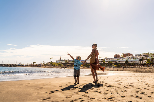 Happy mother and little child holding hands and walking on beach, talking, smiling and showing clouds on the sky.