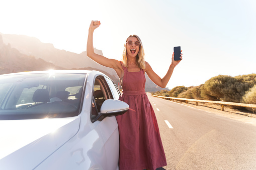 Happy blonde woman with mobile phone in one hand standing near the car, using navigation online to get to her dream destination on holidays. Tourist lifestyle. Real people emotions. Traveler.