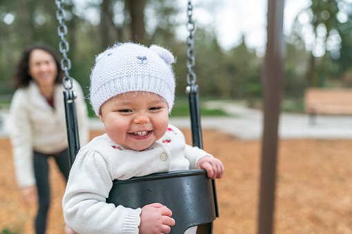 An adorable and happy one year old baby is on a swing at the park while her mother smiles and pushes her from behind. Both the mom and baby are smiling and laughing.