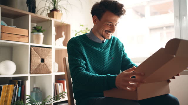 One young happy Caucasian man sitting at home and unpacking a parcel that he order from an online store. Satisfied consumer. Online shopping.