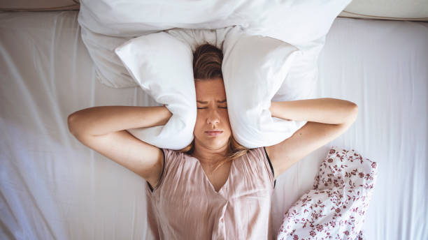 Young woman trying to sleep but disturbed by noisy neighbors and covering ears with pillows Young woman trying to sleep but disturbed by noisy neighbors and covering ears with pillows. I can's sleep with all that noise! Sleep problems. Angry woman disturbed with a noise trying to sleep evil stock pictures, royalty-free photos & images