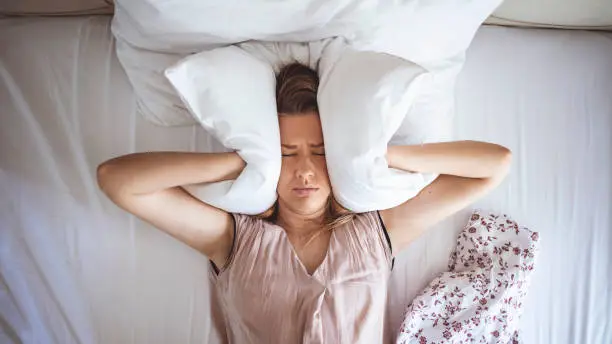 Photo of Young woman trying to sleep but disturbed by noisy neighbors and covering ears with pillows