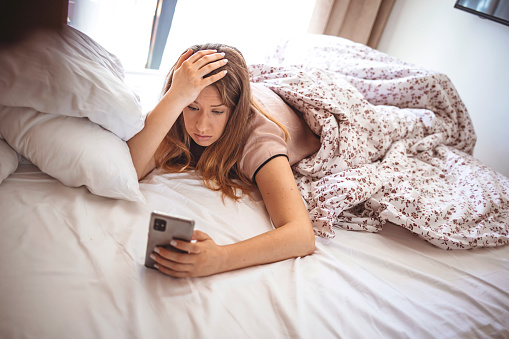 Young woman is waking up and looking at her smart phone. Woman upset after reading bad message on phone at home. Worried, young teenager looking at the phone in her room