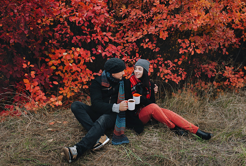 A young couple in love in a coat, in hats and with scarves sits on the grass near the red autumn bushes and drink coffee. The concept of rest and relaxation in nature.