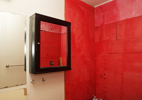 Bright red boards with a waterproofing chemical line a shower wall during a bathtub to shower conversion. The mirror on the medicine cabinet reflects the wall openings for the shower head and controls. Canadian house built in 1987.