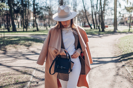 Stylish young woman wears hat holding handbag walking in park. Spring female clothes and accessories. Fashion