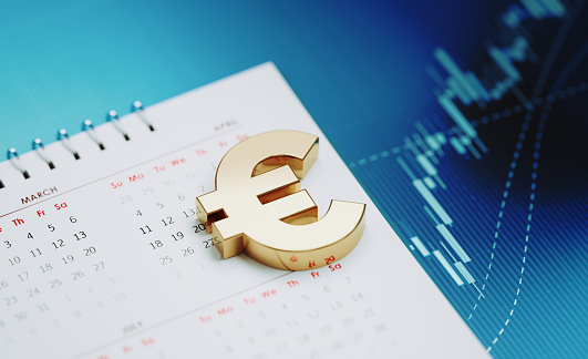 Gold colored Euro symbol sitting over a white calendar on blue financial graph. Horizontal composition with selective focus and copy space. Investment, stock market data and financial planning concept.