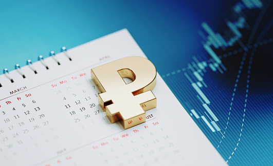 Gold colored Russian Rubles sign sitting over a white calendar on blue financial graph. Horizontal composition with selective focus and copy space. Investment, stock market data and financial planning concept.