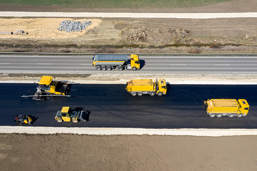 Aerial view of an asphalting paver machine and other construction machinery and vehicles during asphalting works on a new road.