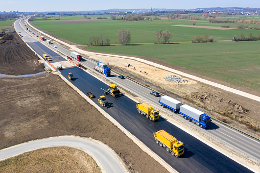 Asphalting paver machine and other modern construction machinery and vehicles during asphalting works on a second lane of a highway, aerial view.
