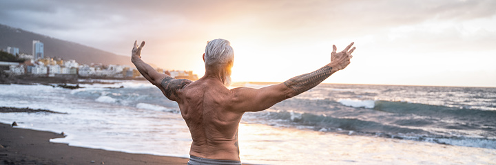 Age is just a number. In a healthy body, healthy mind. Senior man showing his muscular fit body with tattoos on the beach. Summer vacation workout. Elderly people healthy lifestyle on a retirement concept.