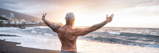 Age is just a number. In a healthy body, healthy mind. Senior man showing his muscular fit body Age is just a number. In a healthy body, healthy mind. Senior man showing his muscular fit body with tattoos on the beach. Summer vacation workout. Elderly people healthy lifestyle on a retirement concept. arm tattoo ideas for men pictures stock pictures, royalty-free photos & images