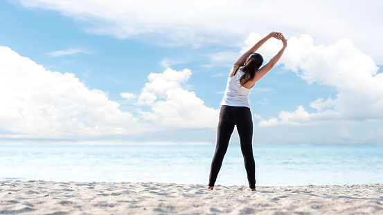 Lifestyle athletic woman warm up and raise arm before yoga exercise for healthy life.  Young girl or people pose balance body vital zen and meditation workout and fitness sport outdoor on sand beach. Sea and sky background. Health care Concept