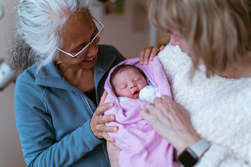 A beautiful moment that shows a mixed race asian grandmother and a Caucasian grandmother holding their newborn granddaughter for the first time in the hospital room. The baby is swaddled and content.