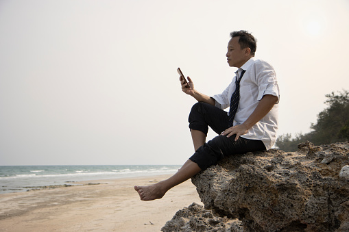 Relax Asian businessman in casual cloth sitting on a cliff at beach with less tense or anxious at the end of day holding mobile smartphone connecting people on social network at the end of day