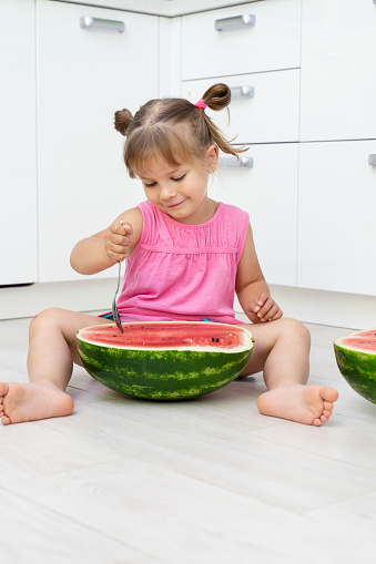 funny little girl eating watermelon with a large spoon while sitting on the floor in a white kitchen