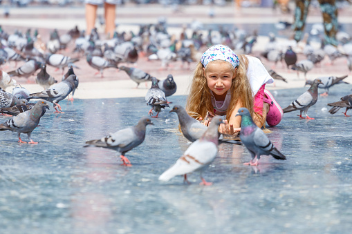 A beautiful Caucasian blonde girl with hair, 6-7 years old, feeding pigeons at Plaza de Catalunya in Barcelona. Bird watching. Summer vacation, travel to Europe. Childhood, carelessness. Real life.