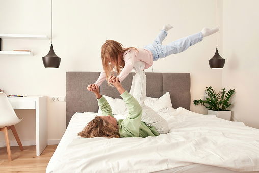 Brother and sister having fun on the bed. Boy is holding his sister on his legs white furniture and a plant on a background