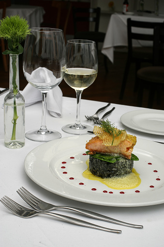 A beautifully plated salmon dish on a bed of spinace. The food is on a white plate with red reduction around it as sauce. Wine glasses and cutlery is also seen in the image.
