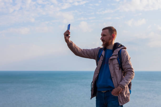 istock Bearded man in raincoat with backpack on sea landscape blue cloudy sky background with smartphone in hand taking picture 1311229197
