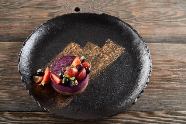From above view of round piece of traditional blueberry cheesecake served with blueberries and strawberries on wooden table in restaurant. Golden brushstroke decoration on black plate.